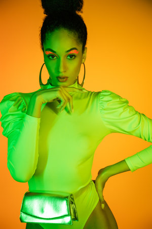 Trendy african american woman in neon bodysuit with waist bag standing isolated on orangeの写真素材