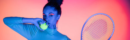 Pretty african american model with neon visage holding ball and tennis racket on pink background, bannerの写真素材
