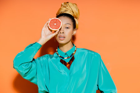 Fashionable african american woman with headscarf holding grapefruit on orange backgroundの写真素材