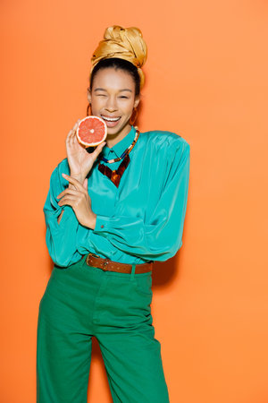 Stylish african american woman in summer clothes holding grapefruit and winking at camera on orange backgroundの写真素材
