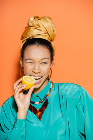 Portrait of stylish african american woman biting lemon on orange backgroundの写真素材