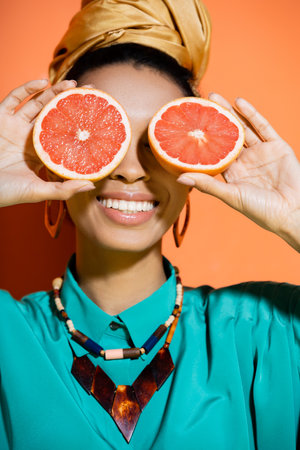 positive and stylish african american model covering face with grapefruit on orange backgroundの写真素材