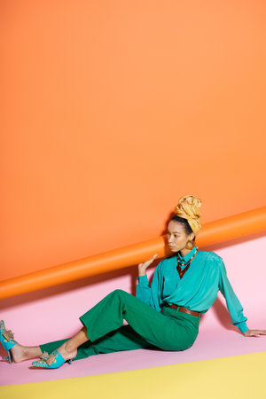 Trendy african american model in bright blouse and headscarf sitting on colorful backgroundの写真素材