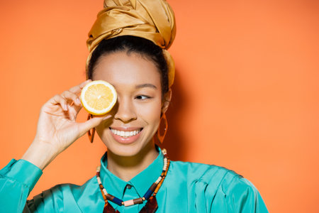Fashionable african american woman smiling and holding lemon on orange backgroundの写真素材