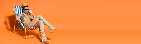Trendy and smiling african american woman in swimwear waving hand while sitting on deck chair on orange background, bannerの写真素材
