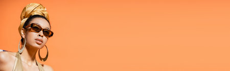 Trendy african american model in golden headscarf and sunglasses isolated on orange, bannerの写真素材