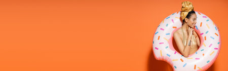 Cheerful african american model in golden headscarf posing with pool ring on orange background, bannerの写真素材
