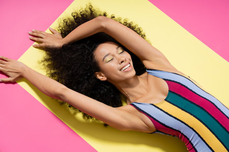 top view of happy african american woman in colorful swimsuit getting tan on yellow and pink backgroundの写真素材