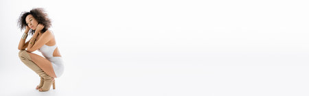 full length of african american model in tight mini dress and knee boots sitting on grey background, bannerの写真素材