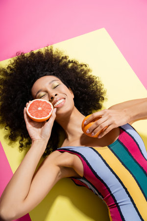 high angle view of pleased african american woman in colorful swimsuit holding grapefruit halves on yellow and pinkの写真素材