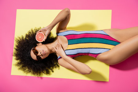 top view of young african american woman in sunglasses and colorful swimsuit holding grapefruit half on yellow backgroundの写真素材