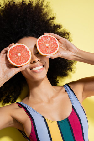 top view of happy african american woman in colorful swimsuit covering eyes with grapefruit halves on yellow backgroundの写真素材