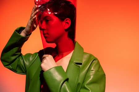 brunette woman in green stylish jacket adjusting transparent cylinder shape plastic on head on orange backgroundの写真素材
