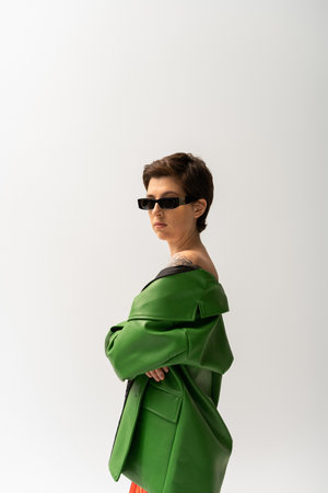 brunette tattooed woman in green leather jacket and stylish sunglasses looking at camera isolated on greyの写真素材