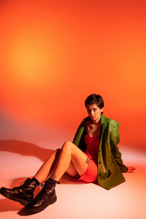 full length of fashionable brunette woman in green jacket and black leather boots sitting on orange backgroundの写真素材