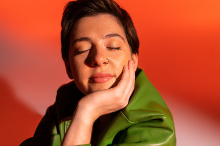 positive and dreamy woman with closed eyes holding hand near face on orange and pink background