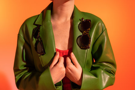partial view of woman with stylish sunglasses on green leather jacket on orange backgroundの写真素材
