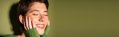 portrait of carefree freckled woman smiling with closed eyes and holding hands near face on green...
