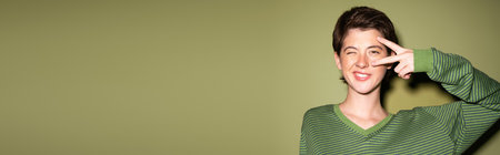 smiling brunette woman winking at camera and showing peace sign near face on green background, bannerの写真素材