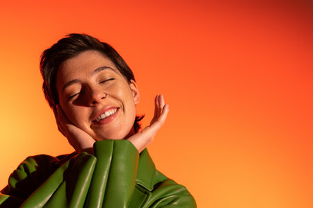 pleased brunette woman in green leather jacket holding hands near face and smiling with closed eyes...