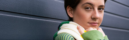 portrait of positive freckled woman looking at camera near grey wall outdoors, banner