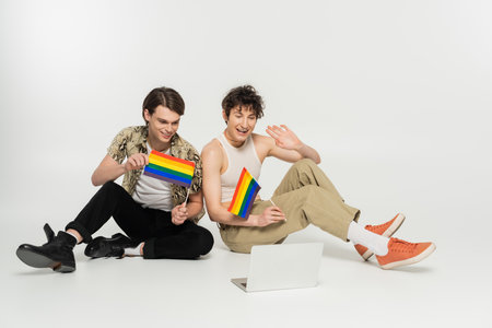happy pansexual couple with lgbt flag having video chat on laptop while sitting on grey backgroundの写真素材