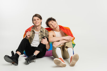 full length of trendy and joyful nonbinary partners sitting with rainbow flag on grey backgroundの写真素材