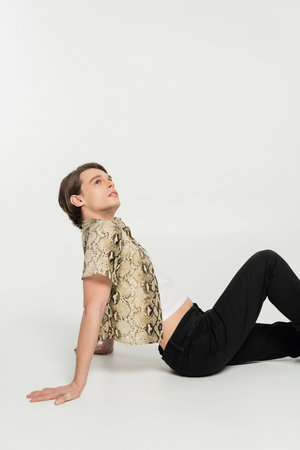 side view of young pansexual person in trendy clothes looking up while sitting on grey background