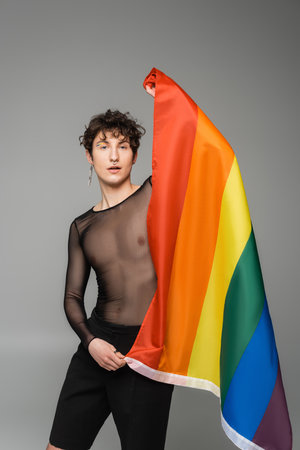 young bigender model in black transparent top holding lgbt flag isolated on greyの写真素材