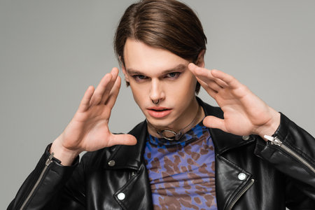 portrait of trendy bigender model in black leather jacket holding hands near face while looking at...