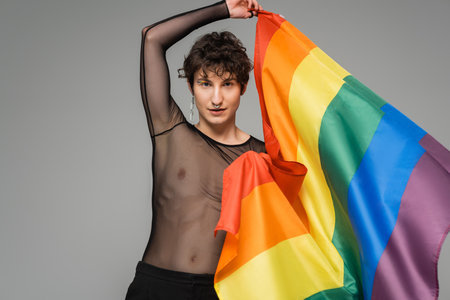 brunette bigender person in black transparent top holding rainbow flag isolated on greyの写真素材