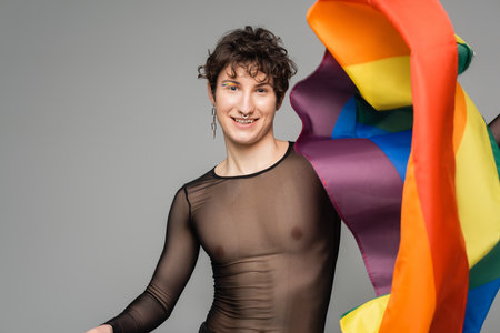 cheerful nonbinary person in black transparent top and silver accessories holding lgbt flag isolated on greyの写真素材