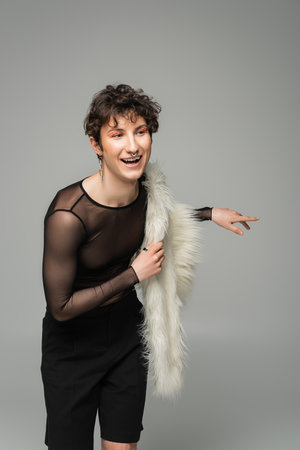 cheerful pangender person with white faux fur jacket looking away and smiling isolated on greyの写真素材