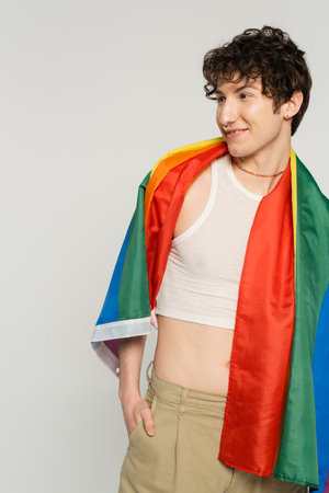 carefree nonbinary person with lgbt flag on shoulders looking away and smiling isolated on greyの写真素材