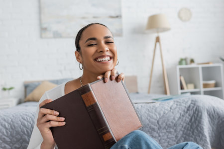 pleased african american student holding book while studying from homeの写真素材