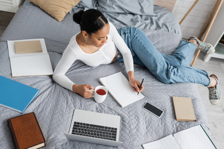 top view of african american student holding cup of tea and writing on notebook in bedroomの写真素材