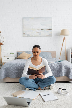young african american student sitting near gadgets and reading bookの写真素材
