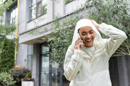 happy african american woman in waterproof raincoat with hood talking on smartphone while standing under rainの写真素材