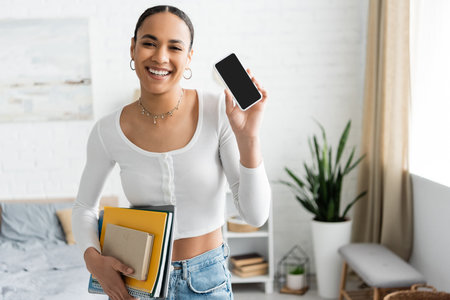 Smiling african american student holding notebooks and showing cellphone in bedroom