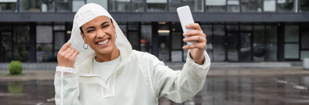 cheerful african american woman in waterproof raincoat taking selfie on smartphone during rain,...