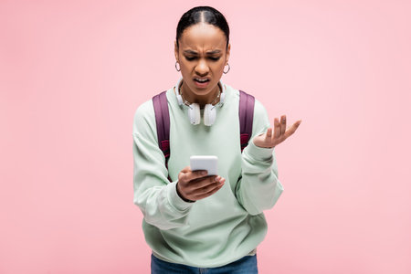 Angry african american student with headphones using mobile phone isolated on pink