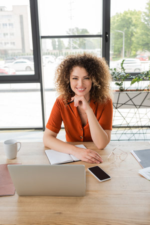 joyful businesswoman with curly hair looking at camera near gadgets and cup of coffee on deskの写真素材