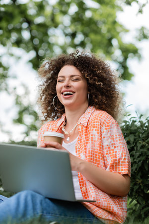 joyful freelancer with curly hair holding paper cup and using laptop outdoors