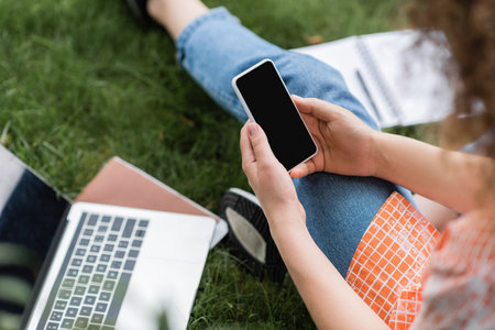 cropped view of young woman holding smartphone with blank screen while sitting near laptop and...