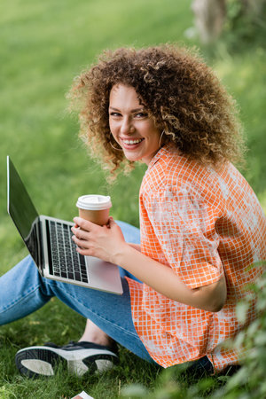 overjoyed freelancer with curly hair holding paper cup and using laptop while sitting on grass