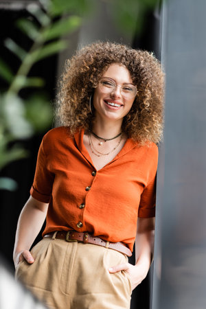 cheerful manager in glasses and hoop earrings standing with hands in pockets near wall in modern...