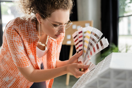 curly architectural designer in glasses holding color palette near residential house model in officeの写真素材
