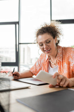 happy designer using graphic tablet and looking at smartphone near laptop while working in officeの写真素材