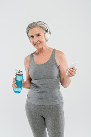 cheerful senior woman listening music in wireless headphones while holding sports bottle and smartphone isolated on greyの写真素材