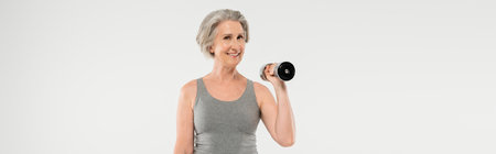 cheerful senior woman with grey hair exercising with dumbbells isolated on grey, bannerの写真素材